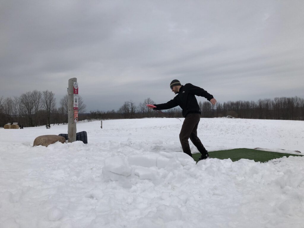 man putting a disc into a disc golf basket in winter