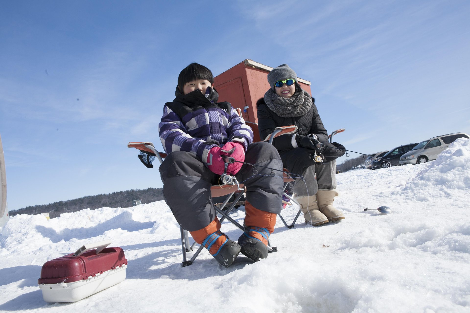 Boy sitting on a chair fishing by a hole in the ice on Rice Lake with another person and an ice hut behind