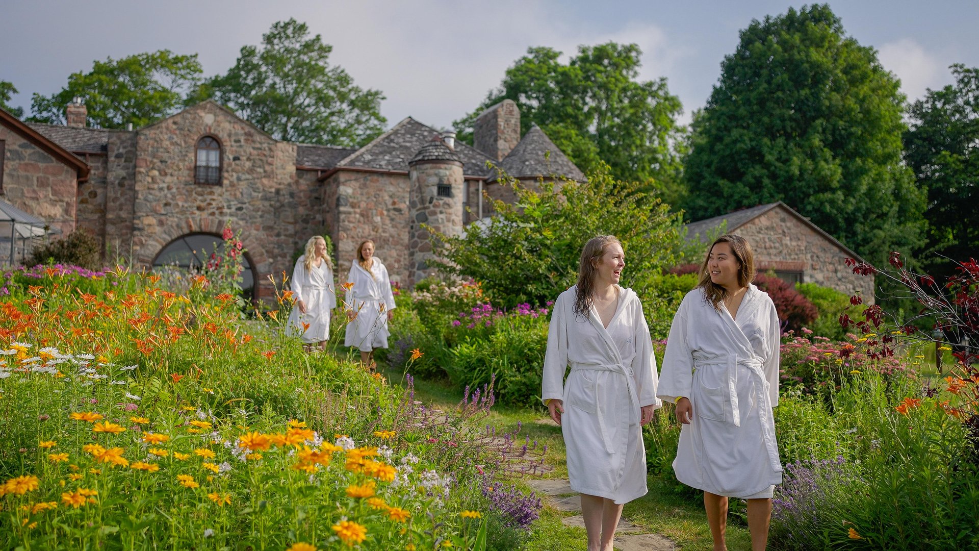 4 women walking a path between gardens with a stone building in the background