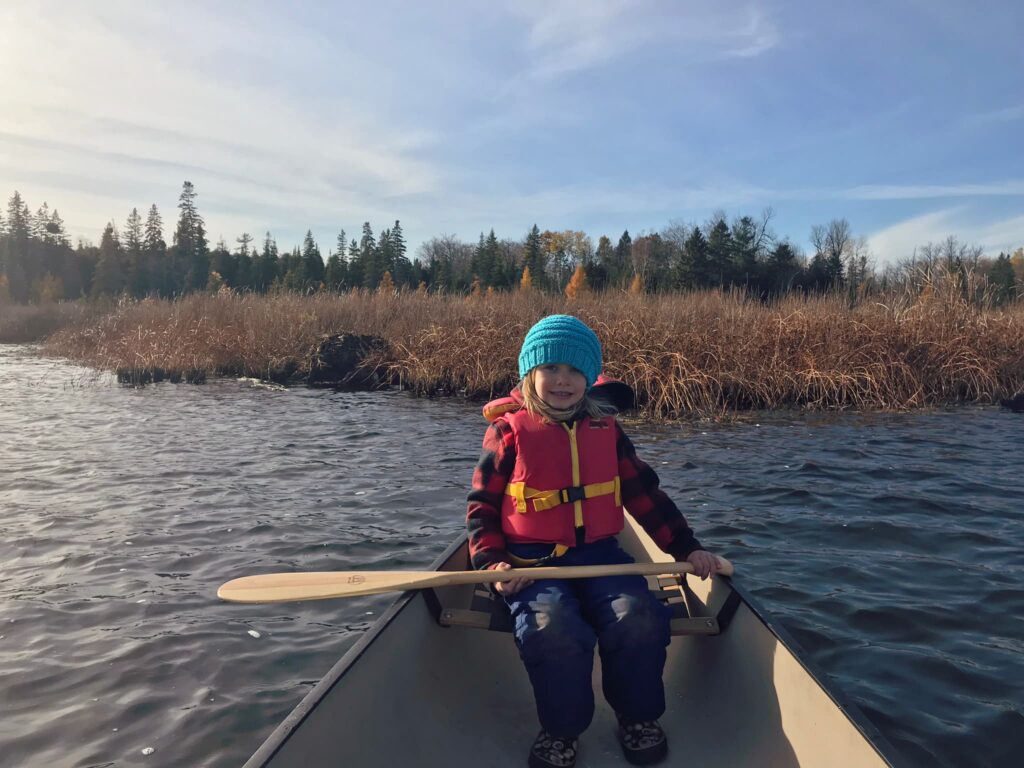 Child with blue hat and red life jacket holding an oar while sitting in a canoe on a lake in fall.