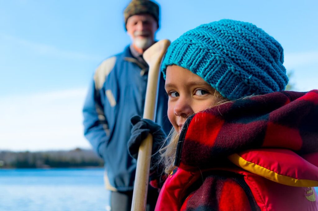 Child with blue hat and red life jacket looks at camera with man looking on in background
