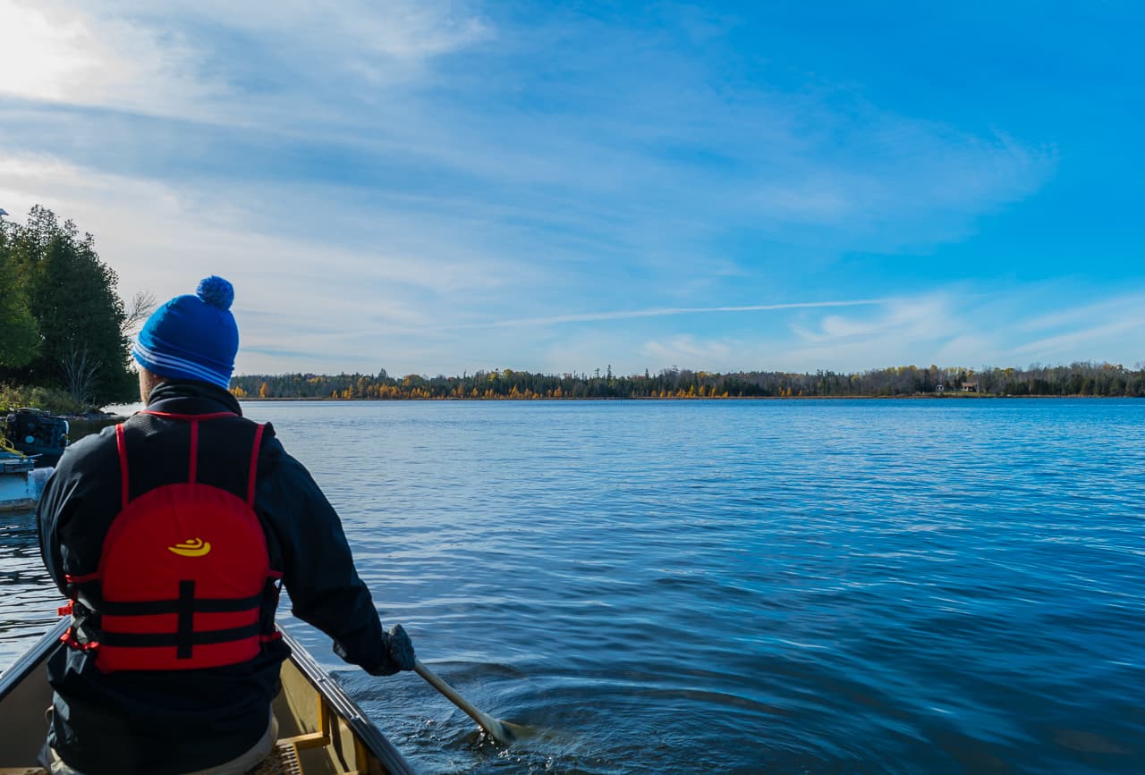 Man with a blue toque on paddling in a canoe on a blue lake in fall