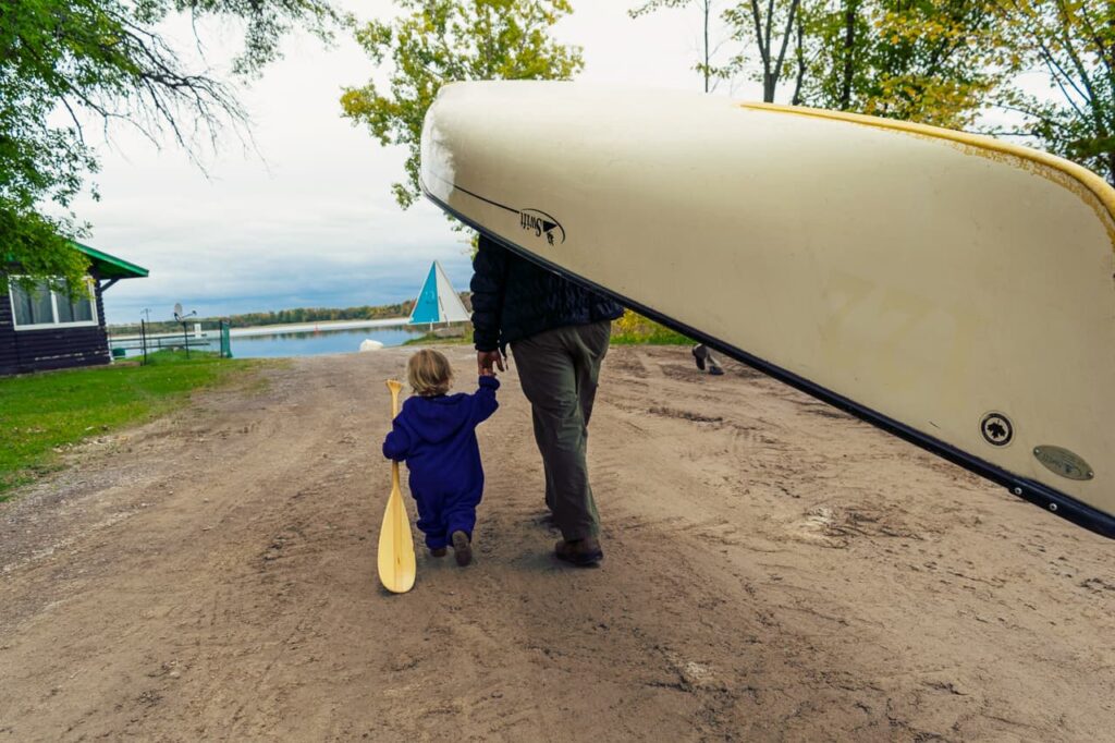 Man portaging a canoe while holding a toddler's hand. Toddler is carrying an oar.