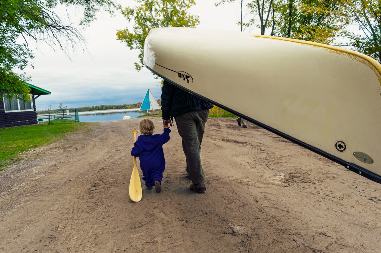 Man portaging a canoe while holding a toddler's hand. Toddler is carrying an oar.