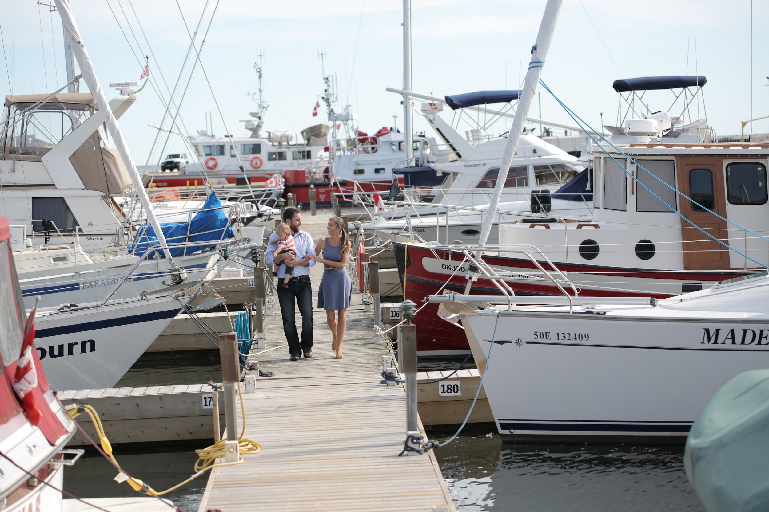 Couple holding a child walking down a slip between sailboats in Cobourg Marina.
