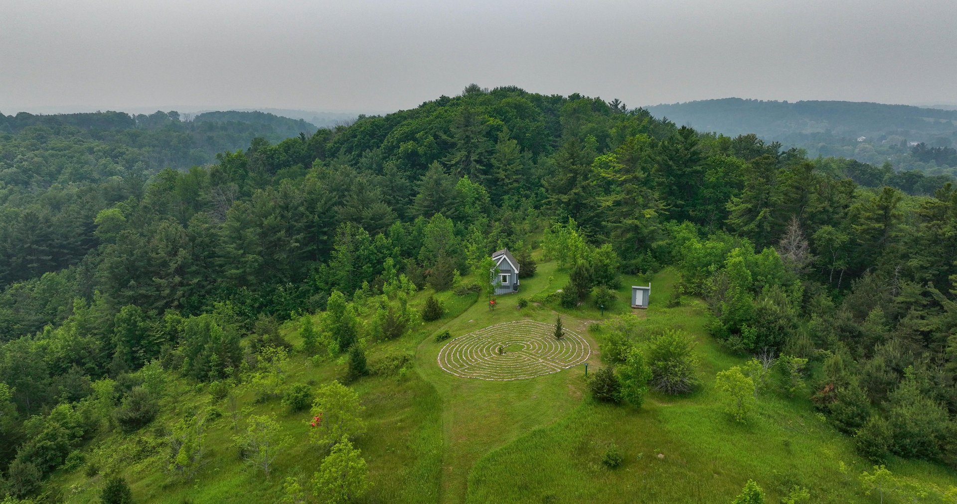 Drone view of Fells Meadows, a lush green hillside with a tiny cabin and a labyrinth