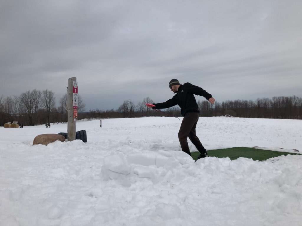 Man throwing a disc towards a disc golf basket in winter