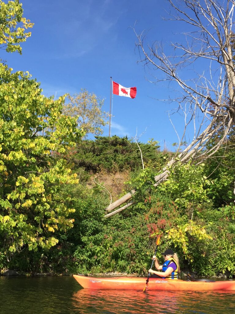 Woman in an orange kayak paddling past a view with a Canadian flag in the background.
