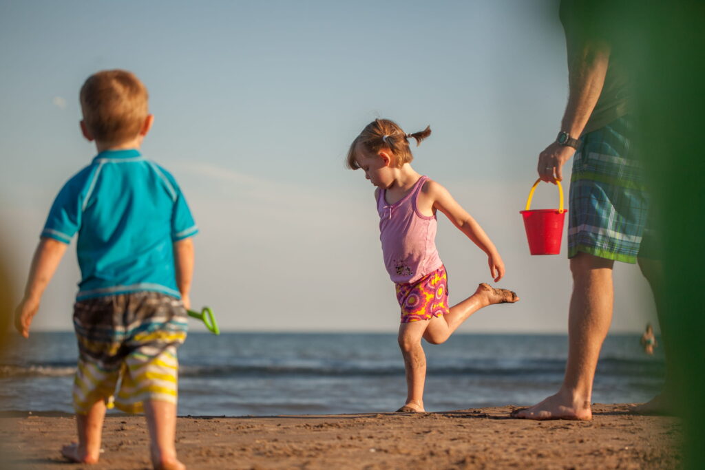 Young girl running on the beach with a boy in front and a man carrying a red bucket.