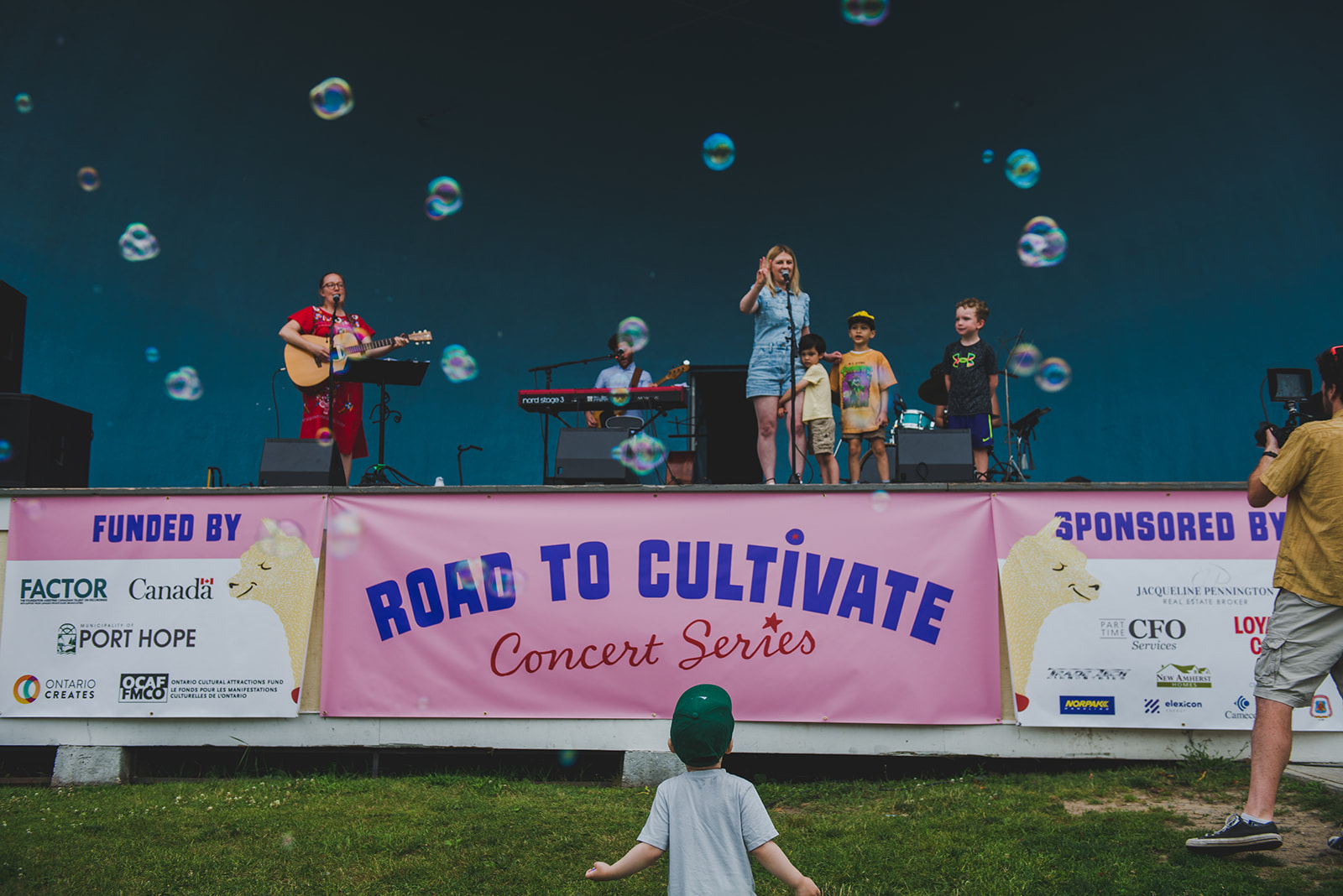 Band playing on an outdoor stage with a sign that reads Road to Cultivate on the stage and a child dancing in the foreground