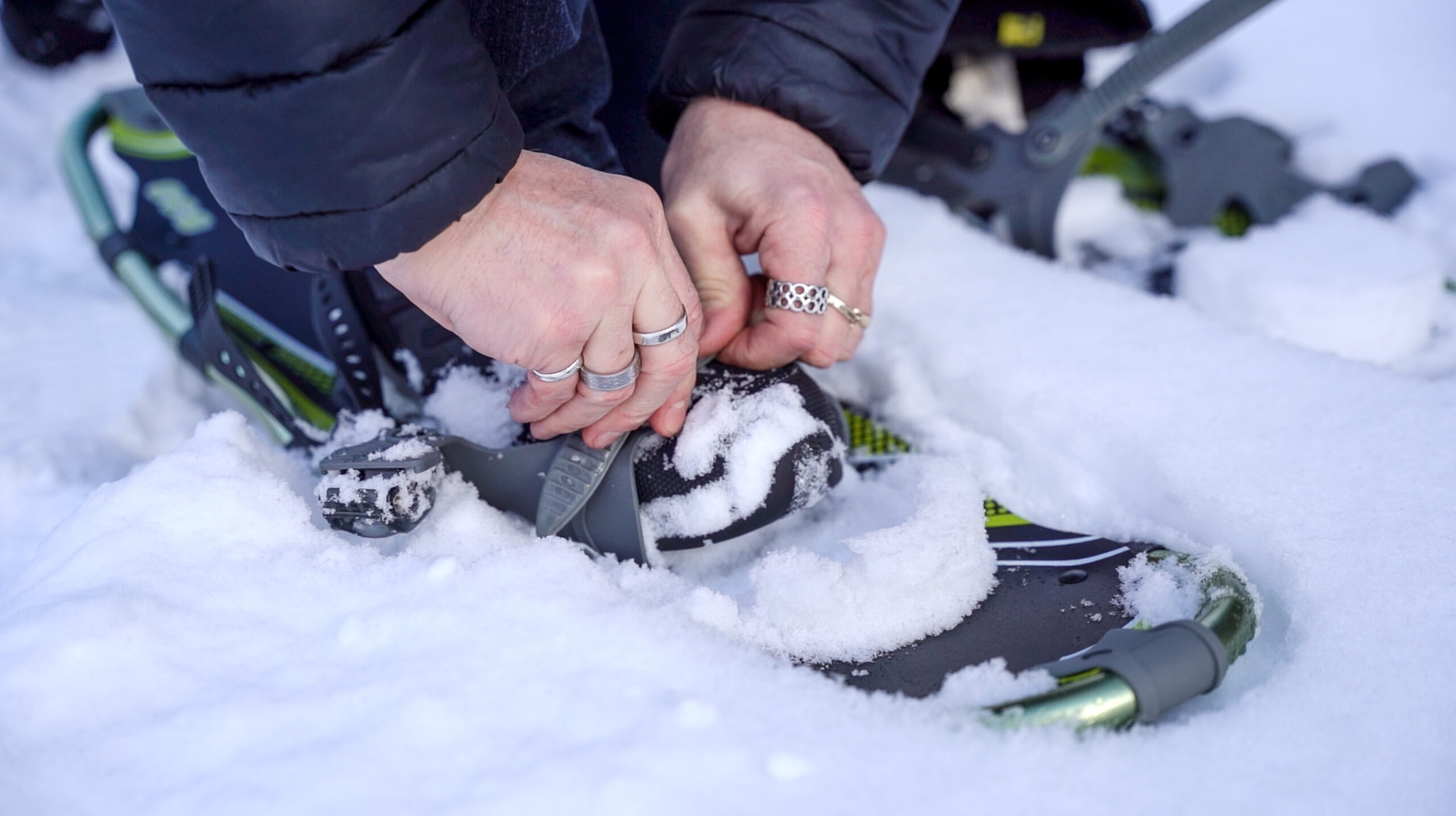 Man's hands wearing multiple silver rings doing up snowshoes.
