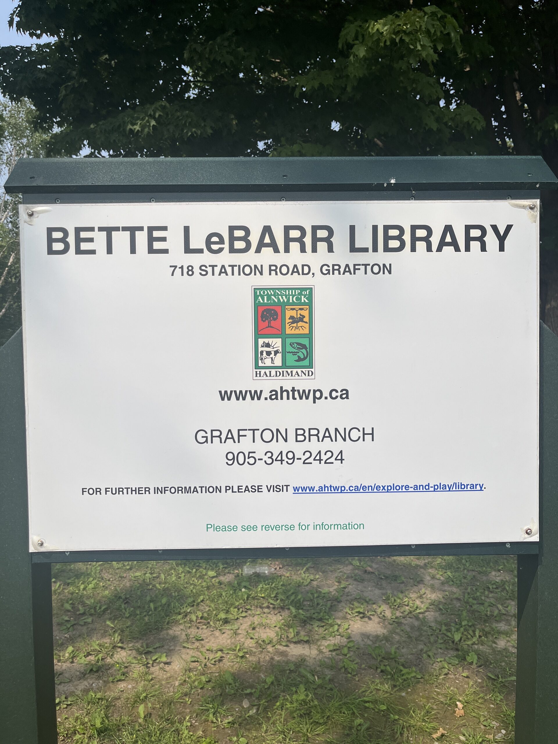 Photo of sign reading "Bette LeBarr Library. 718 Station Road, Grafton. www.ahtwp.ca. Grafton Branch, 905-349-2424. For further information please visit www.ahtwp.ca/en/explore-and-play/library." The Township of Alnwick Haldimand logo is in the middle of the sign.