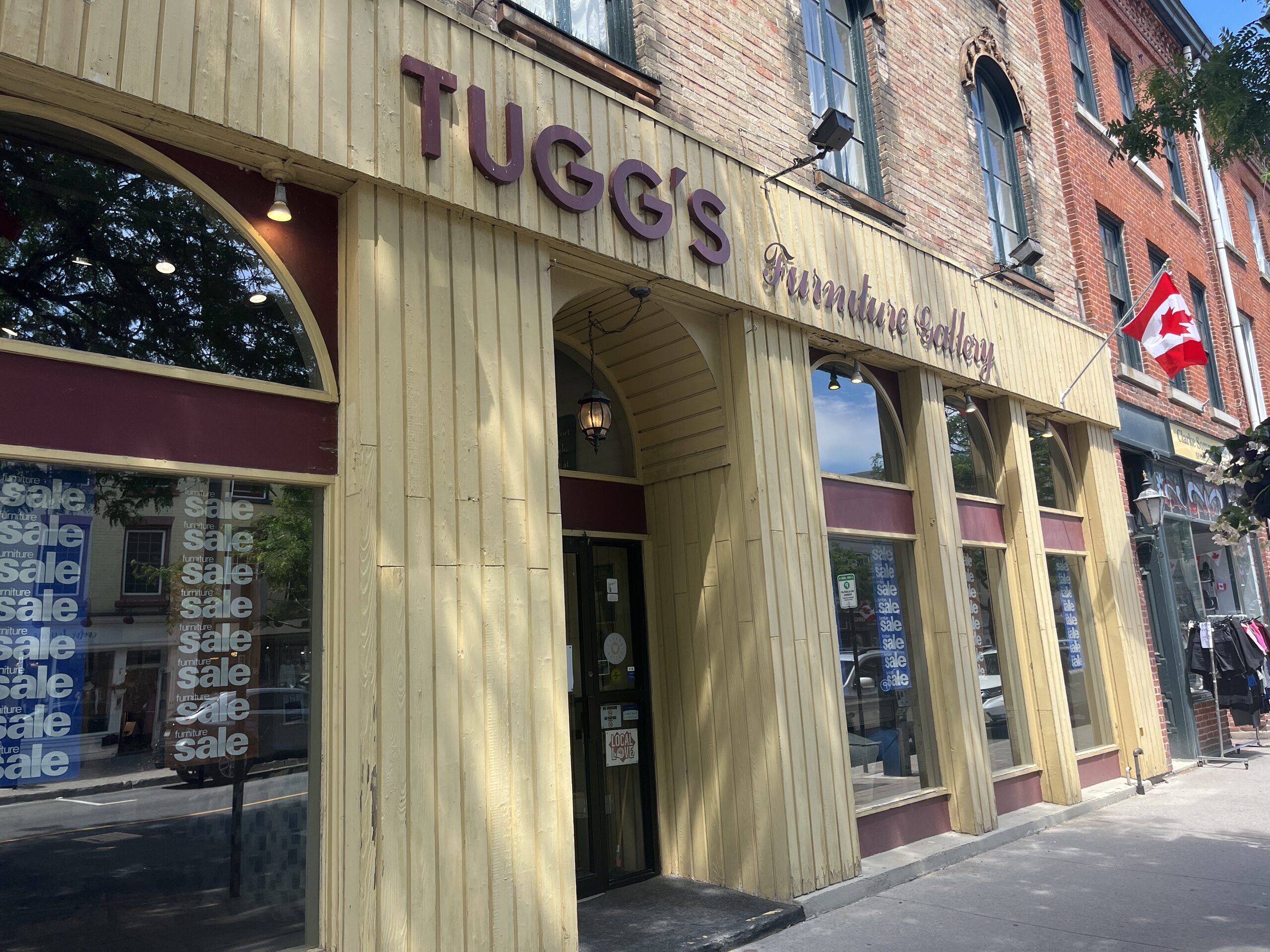 Photo of Tugg's storefront. Yellow paneling with red trim and "Tugg's Furniture Gallery" sign above the door.