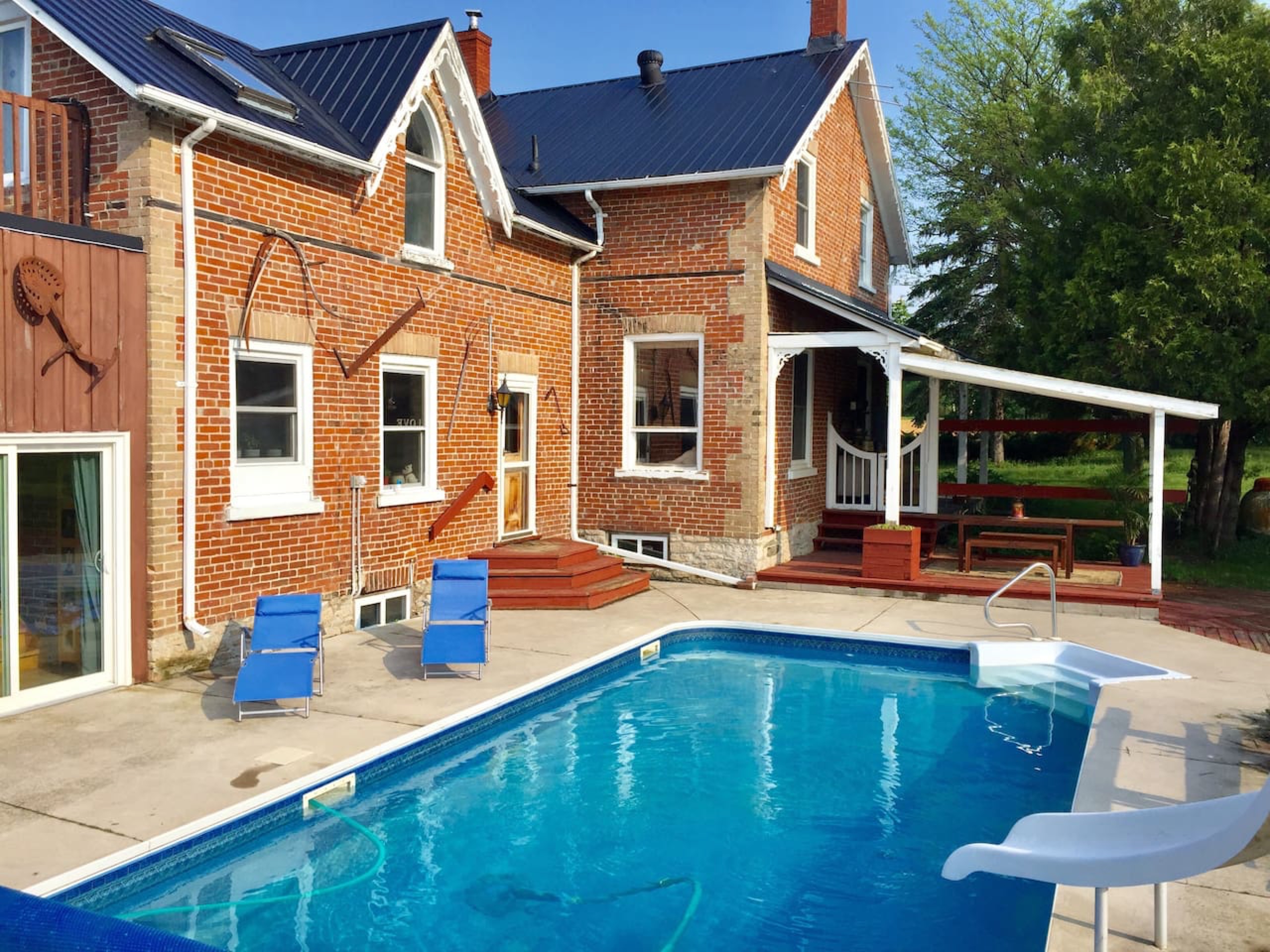 Square in ground pool with a slide and 2 blue lounging chairs in front of a brick house