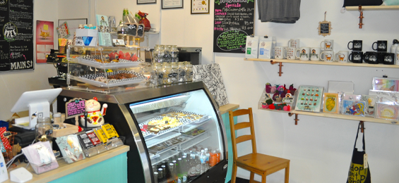 Interior of a cozy café featuring a glass display case filled with pastries and bottled drinks, alongside a counter with a register and assorted treats. Shelves on the wall showcase mugs, cards, and small gift items, adding a boutique charm to the space.