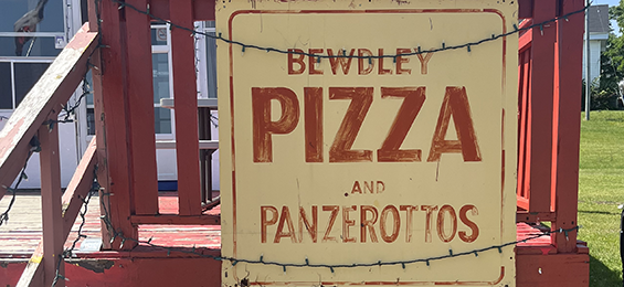 Beige sign with red writing on a red deck with string lights above.