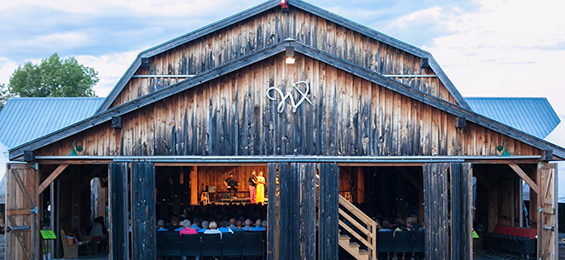 Barn full of people on chairs watching a stage performance at the other end of the barn.