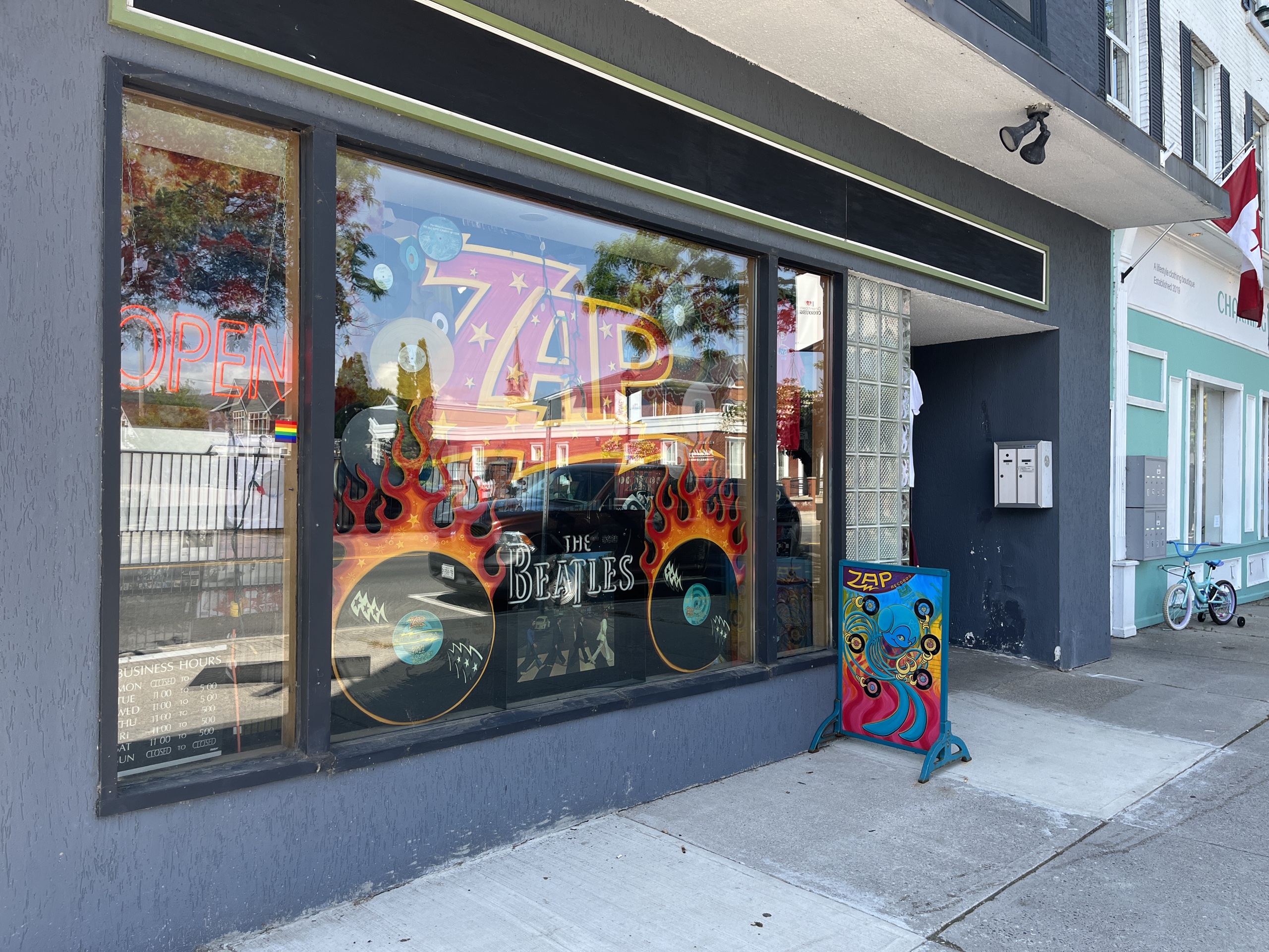 A vibrant storefront for “ZAP” features bold window art, a neon “OPEN” sign, and a colorful sandwich board on the sidewalk.