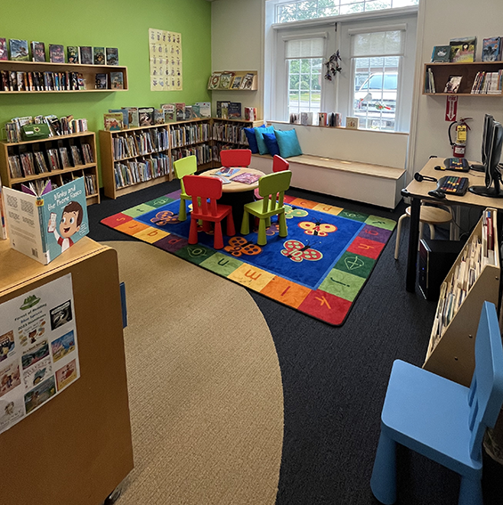 large window seat with pillows and book shelving surrounding a small table wit red and green chairs on an alphabet carpet.