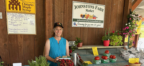 Woman standing holding a flat of strawberries beside a table of fresh vegetables.