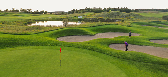 2 people golfing near the greens of a golf hole with 2 bunkers and a pond.