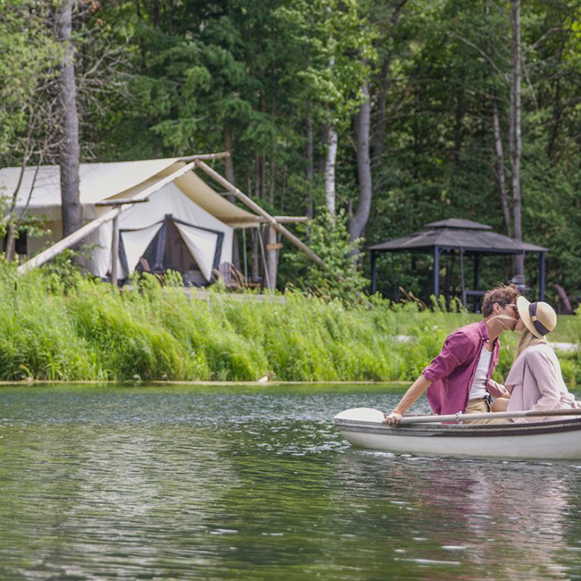 2 people kissing in a canoe near the shore in front of a tent.