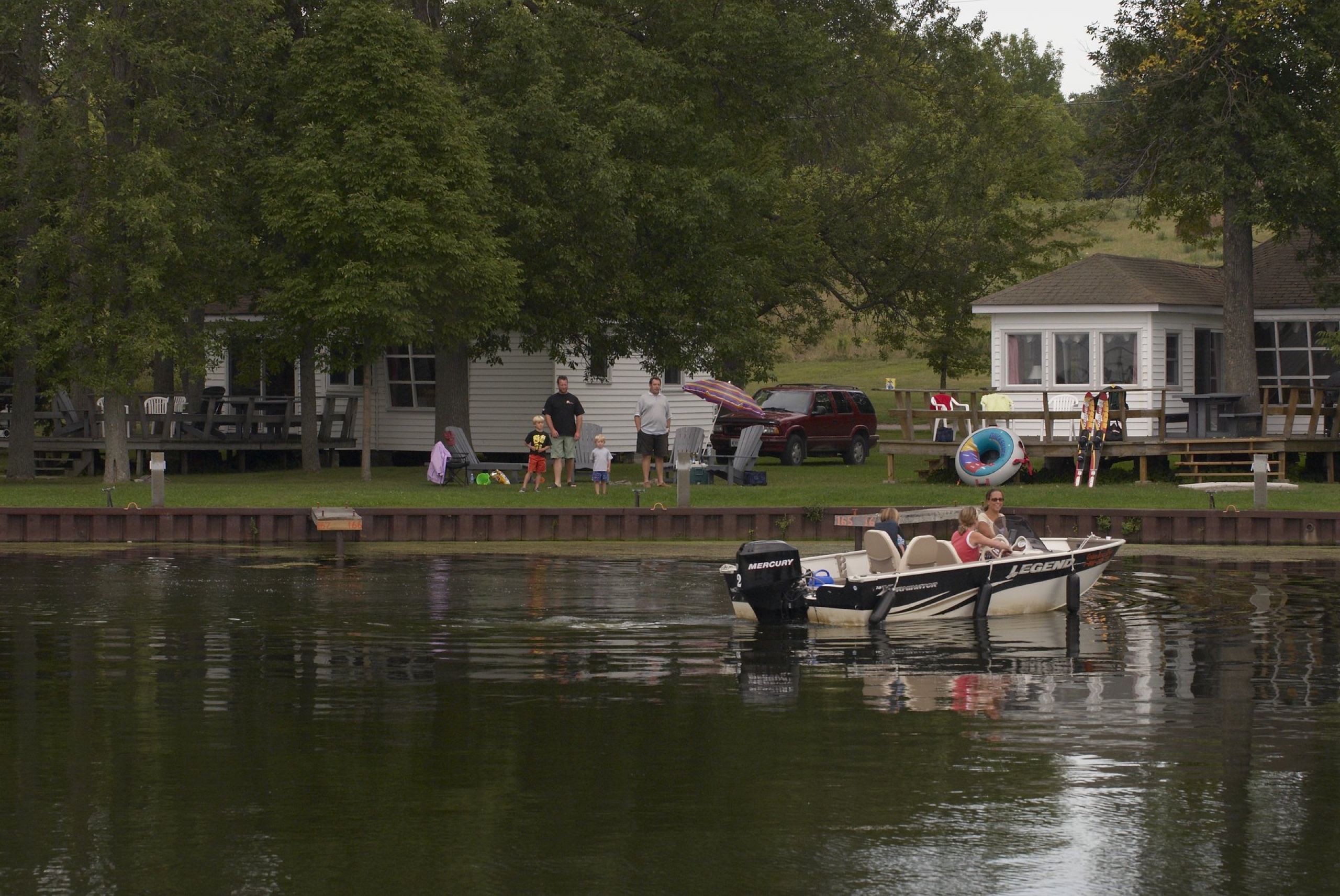 Photo of a boat and cottages on the waterfront. Two fathers and two sons are on the waterfront watching three girls in a boat.