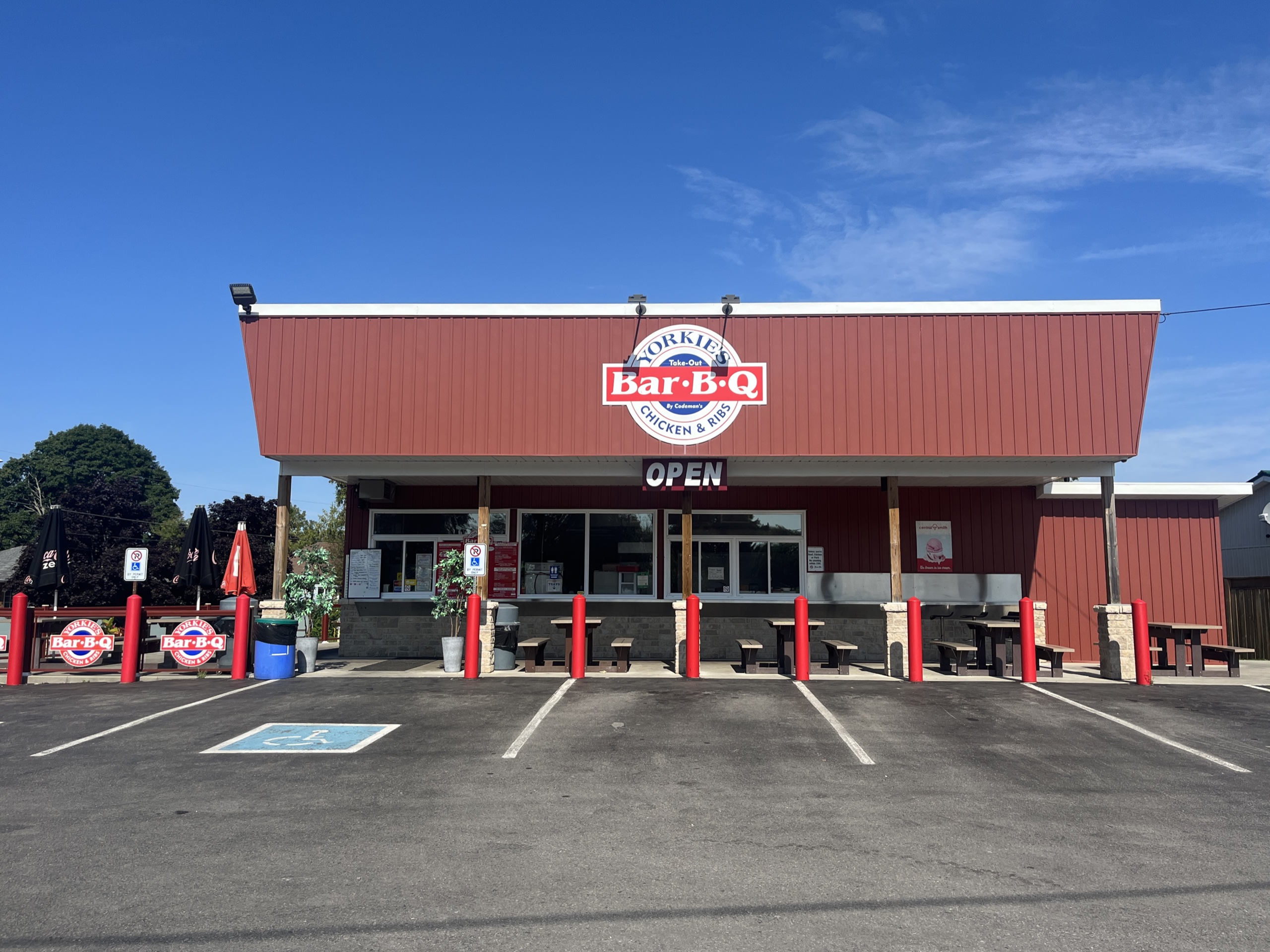 A red and white take-out barbecue restaurant named "Yorkie's Bar-B-Q" with outdoor seating, parking spaces, and a large "OPEN" sign under a clear blue sky.
