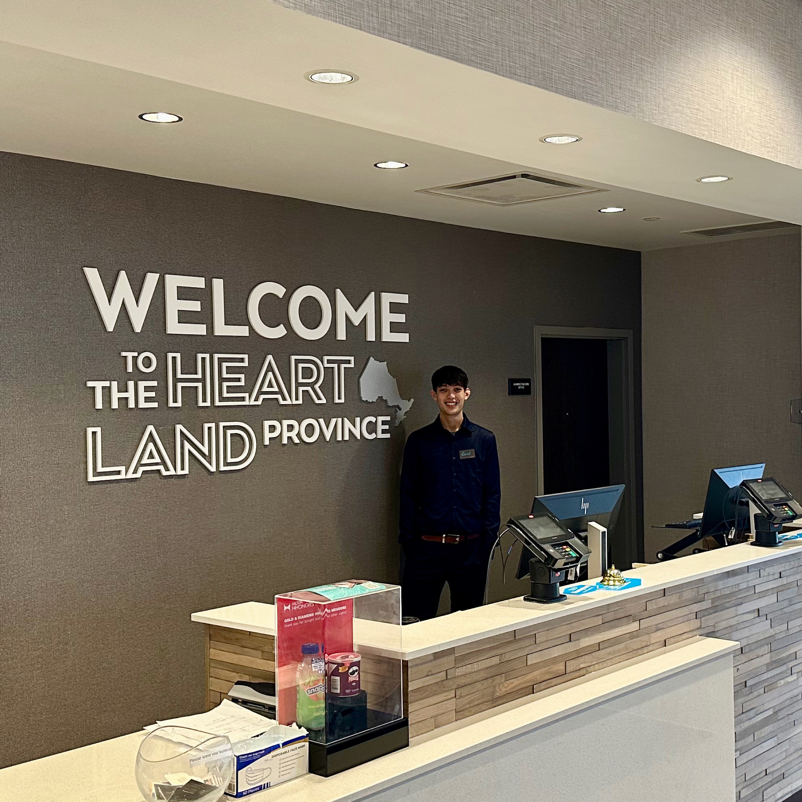 Photo of inside of Hampton Inn Port Hope. Photo is of the front desk with an attendant smiling. On the wall reads "Welcome to the heart land province."