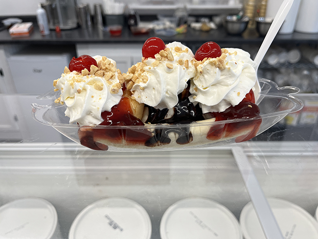 A banana split ice cream treat with 3 piles of whipped cream, sauce, peanuts and 3 cherries.