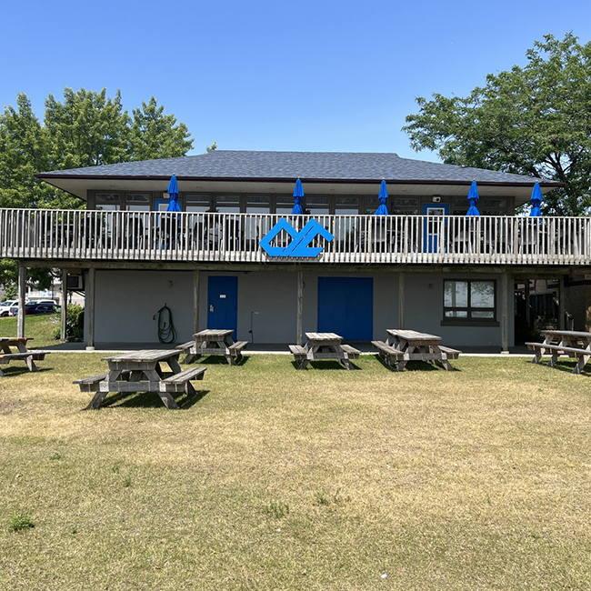 Grey building with a large deck and many picnic tables surrounding.