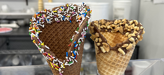 A chocolate waffle cone with sprinkles standing beside a vanilla waffle cone with chocolate and peanuts.