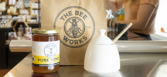 Jar of honey beside a white honeypot and a brown paper bag.