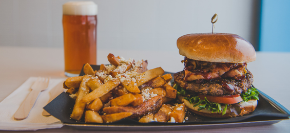 Plate with a large burger and Greek fries in front of a glass of beer.