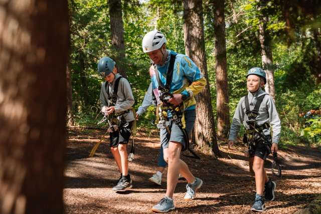Man and two kids with treetop trekking gear on, walking through the forest on a sunny day.