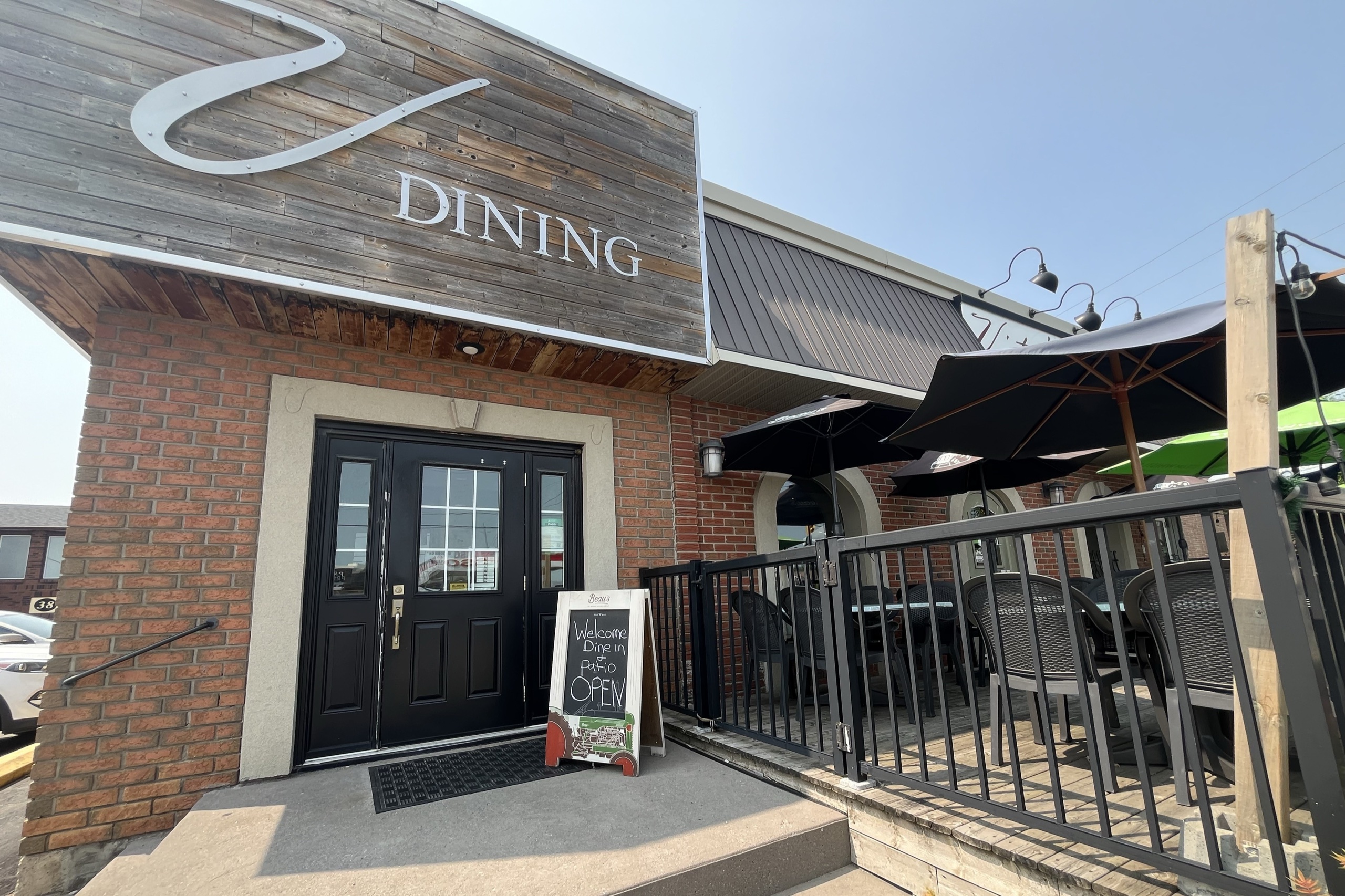 Photo of "Vitos" storefront. There is a red brick building, with a black door and a wooden sign that reads "V Dining". To the right of the door, there is a patio with black umbrellas.