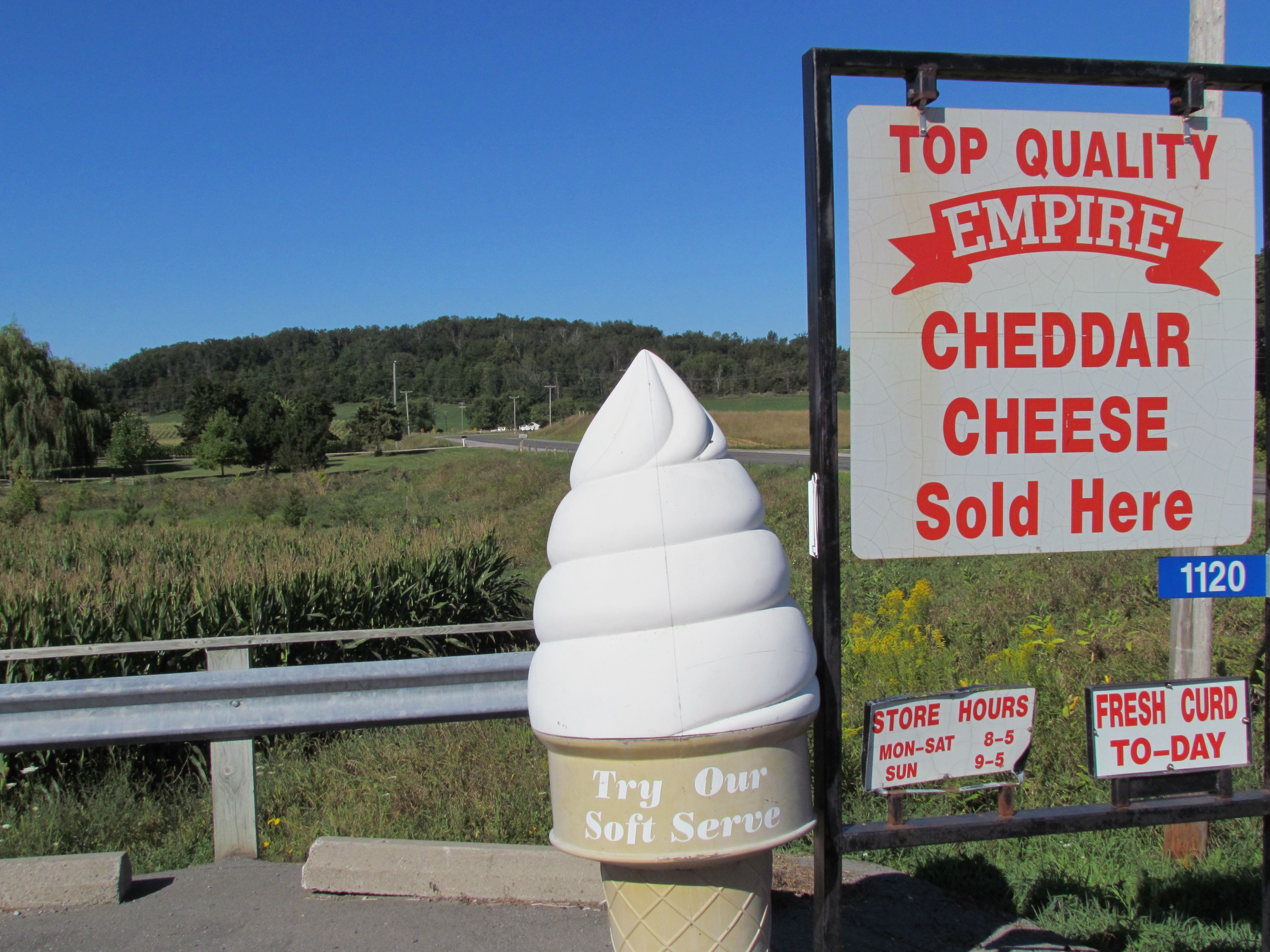Photo of Empire Cheese Co-operative. Photo features ice cream cone and sign. Sign reads "Top quality Empire cheddar cheese sold here. Store hours, Monday to saturday 8-5. Sunday 9-5. Try our soft serve."