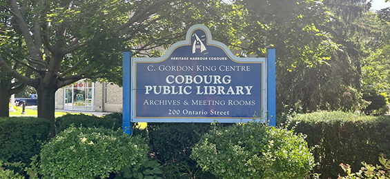 Blue city sign with white writing surrounded by bushes.