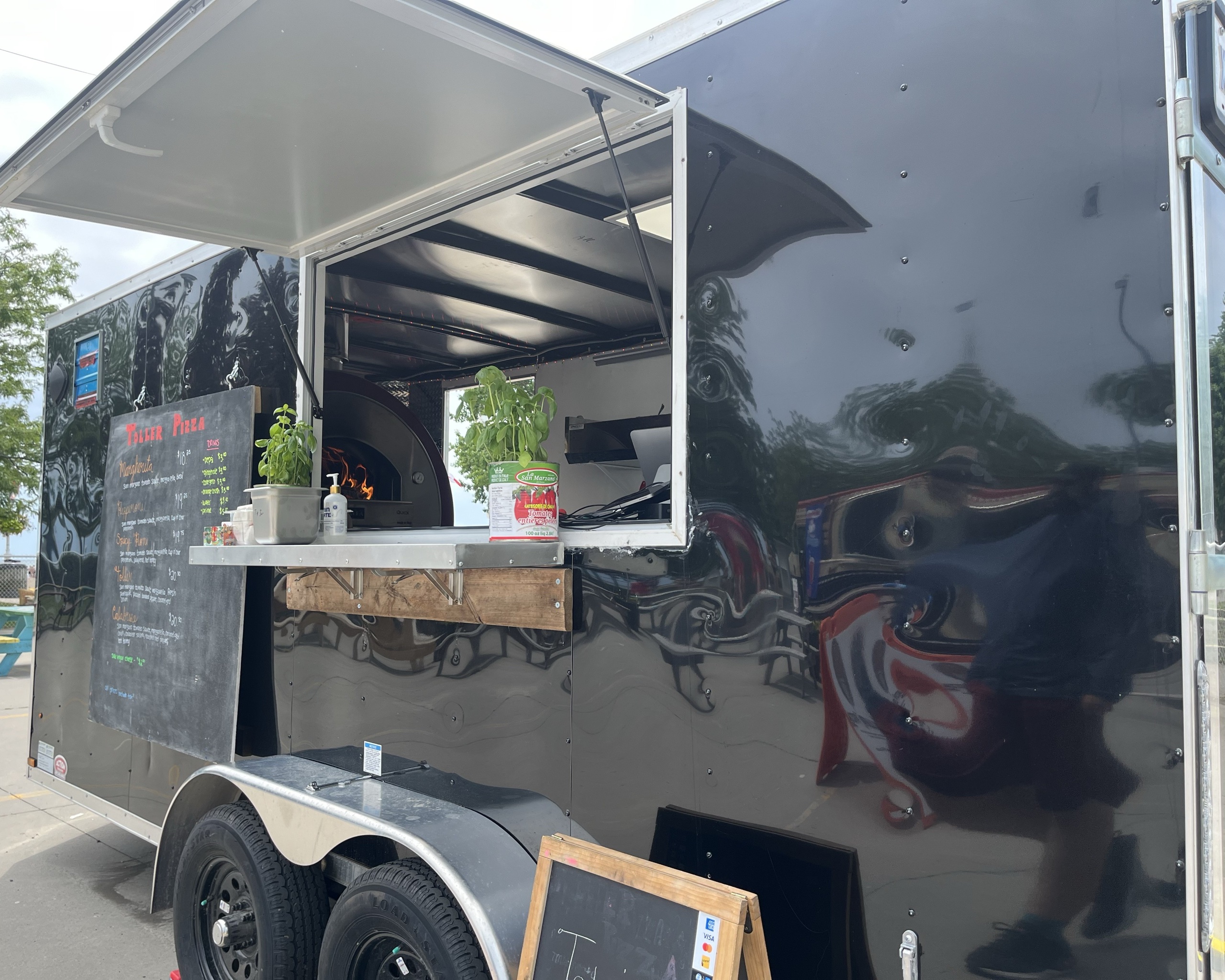 A sleek black food truck with an open service window displays potted basil plants and a can of tomatoes, suggesting fresh ingredients. A wood-fired pizza oven glows inside, with a chalkboard menu listing various pizzas mounted beside the window.