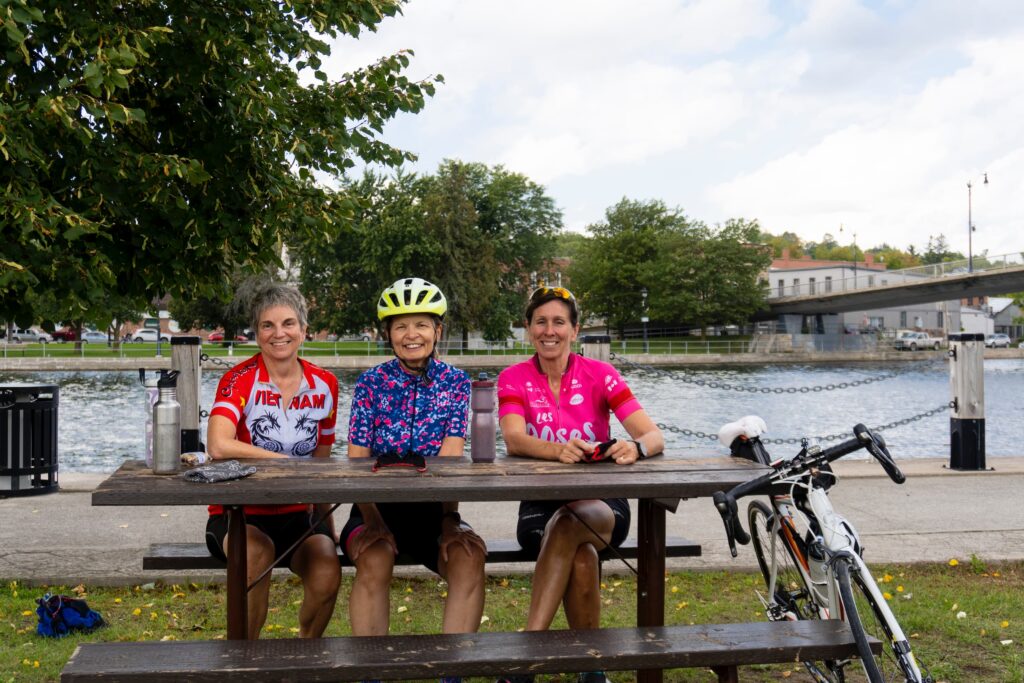 Group of three women smiling at the camera, a bike leans against the picnic table and one woman is wearing a bike helmet