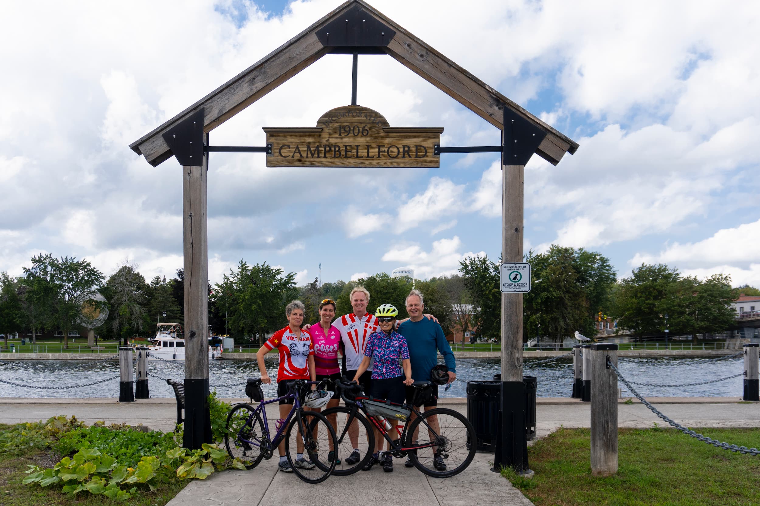 Group of cyclists pose in front of a large Campbellford sign