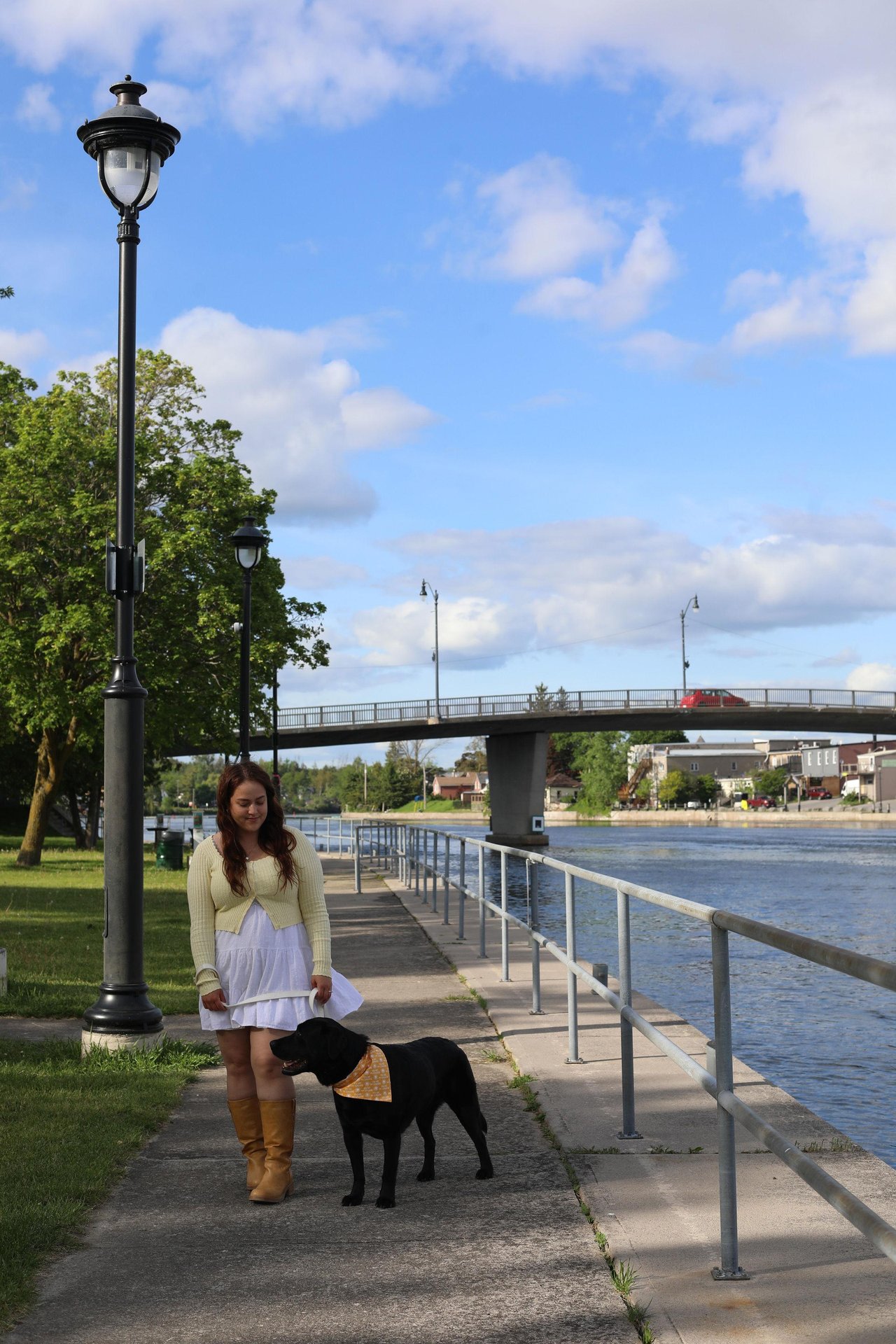 Woman and black lab walking along river's edge with bridge in background