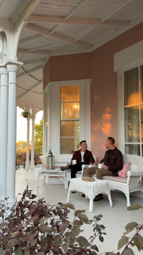 2 men sitting on a porch of a pink Italianate style bed and breakfast.